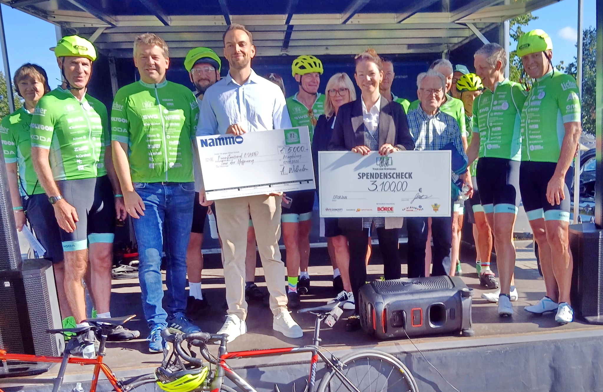 Übergabe von Spendenschecks an die Tour der Hoffnung durch Unterstützer und Sponsoren, umringt von Fahrerinnen und Fahrern in grünen Trikots