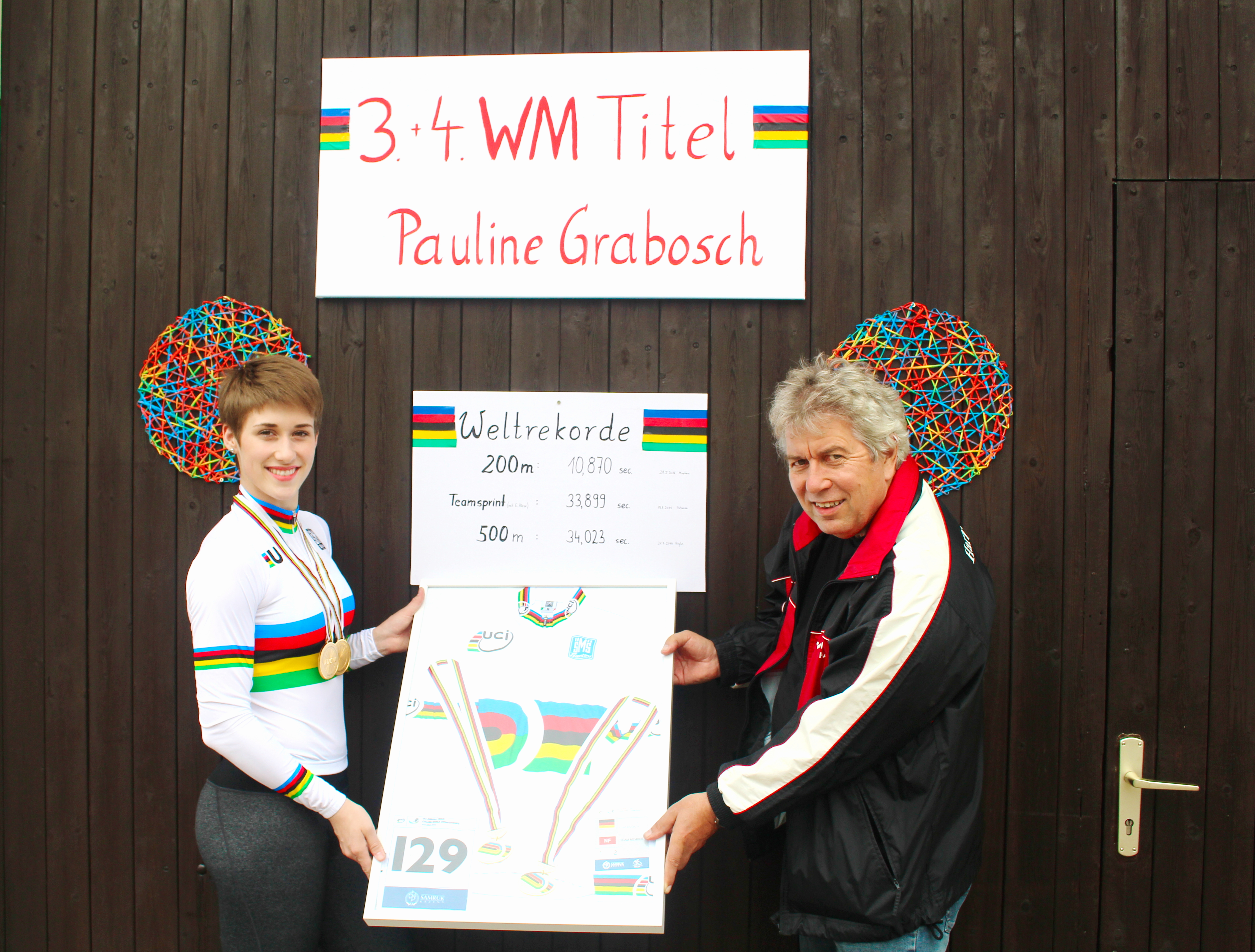 Pauline Grabosch und ein Begleiter präsentieren ein gerahmtes Erinnerungsstück vor einer Tafel mit ihren Weltrekorden